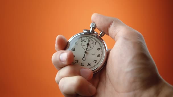 Male hand holding analogue stopwatch on orange color background. Time start with old chronometer man presses start button in the sport concept. Time management concept. - Footage, Video