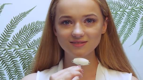 Bella ragazza in foglie tropicali in studio si tocca il viso con un rullo - Filmati, video
