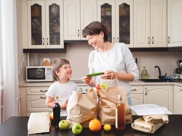 Woman and toddler boys sorts out purchases in the kitchen.  Grocery delivery in paper bags. Subscription service from grocery store. Mother and son at kitchen. - Foto, Bild