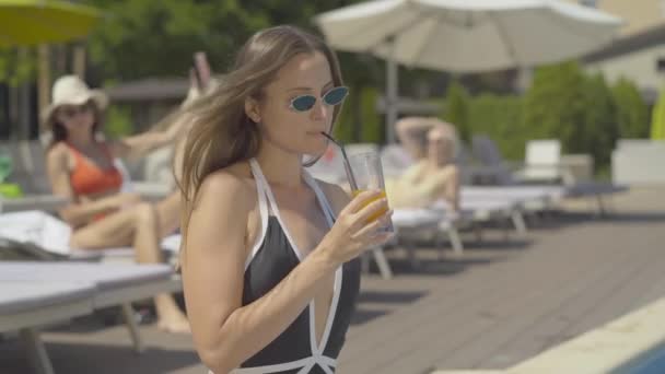 Cheerful brunette woman with cocktail kissing unicorn swimming ring on sunny day. Side view portrait of joyful Caucasian tourist resting at summer resort. Tourism concept. - Filmagem, Vídeo