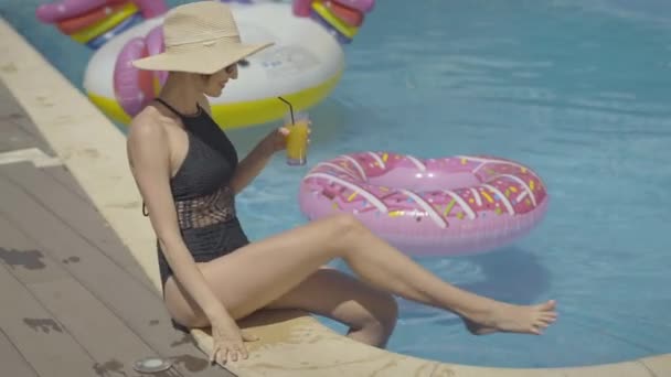 Portrait of happy wealthy woman with cocktail glass having fun at poolside. Smiling adult Caucasian tourist drinking alcohol and splashing water in swimming pool at luxurious resort. - Filmmaterial, Video