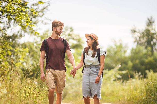 Couple Hiking Along Woodland Path. Happy loving man and woman on holiday walking together. Active lifestyle concept. Young people walking with backpacks on country road outdoors. - Photo, Image