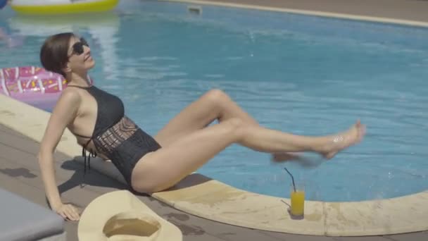 Side view of cheerful brunette woman having fun sitting on poolside. Portrait of beautiful Caucasian lady splashing water and smiling. Happy tourist enjoying vacations on sunny summer day at resort. - Materiaali, video