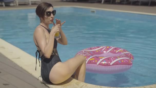 Slim sensual Caucasian woman in sunglasses drinking cocktail and splashing water in swimming pool on sunny day. Portrait of brunette beautiful tourist resting at luxurious resort. - Filmagem, Vídeo