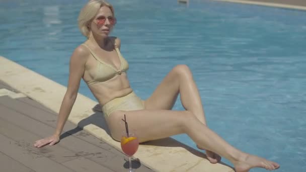 Confident tanned blond woman in golden swimming suit sunbathing at luxurious resort. Wide shot portrait of gorgeous adult Caucasian tourist in sunglasses sitting at poolside on sunny summer day. - Filmagem, Vídeo