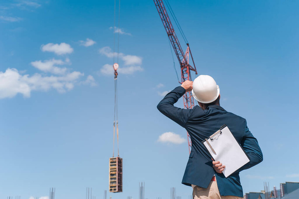 The chief in a jacket and a construction helmet with documents in his hands, against the background of a tower crane and the sky with clouds. House construction inspection - Fotó, kép