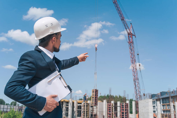 The chief in a jacket and a construction helmet with documents in his hands, against the background of a tower crane and the sky with clouds. House construction inspection - Φωτογραφία, εικόνα