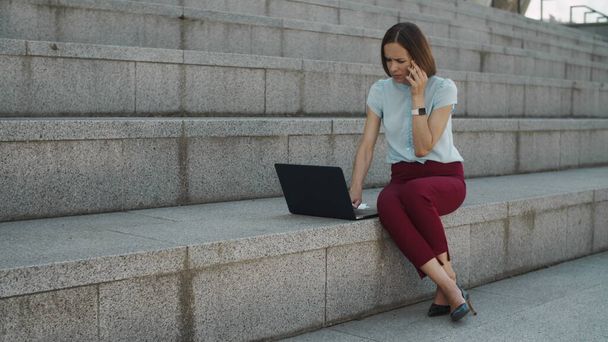 Επιχειρηματίας μιλάει σε smartphone σε εξωτερικούς χώρους. Γυναίκα που εργάζεται σε φορητό υπολογιστή - Φωτογραφία, εικόνα