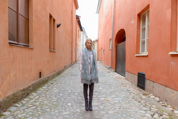 Portrait of young beautiful blonde Scandinavian woman along the alley with orange building in the city - Photo, Image