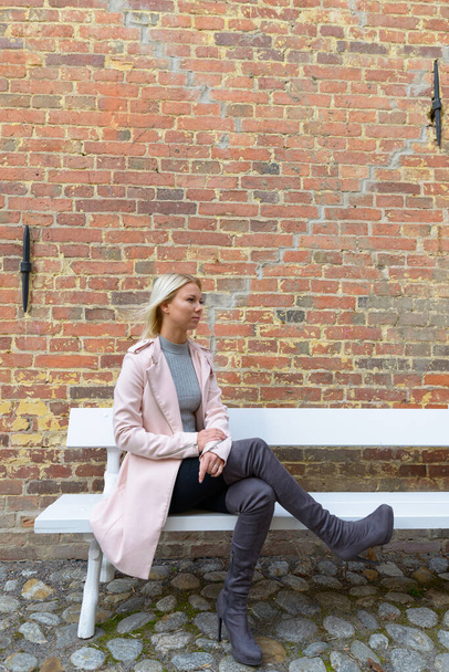 Portrait of young beautiful blonde Scandinavian woman sitting on park bench in the city streets outdoors - Photo, Image
