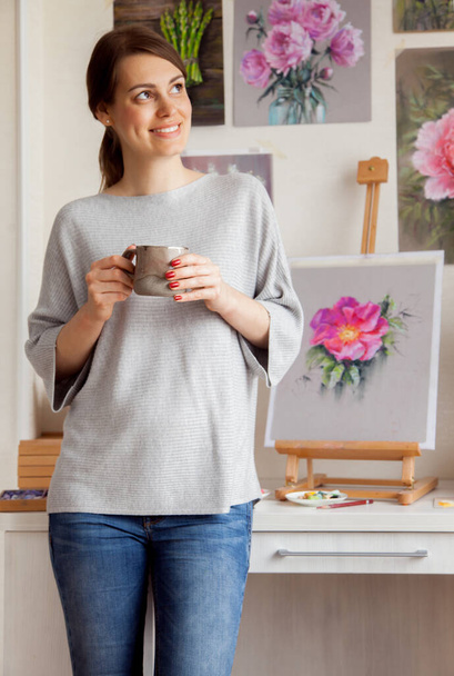 Pretty young barefoot girl artist in casual clothes drinks coffee while relaxing after writing a sketch of a bright pink rose hip standing on a table on an easel and tools - Fotó, kép