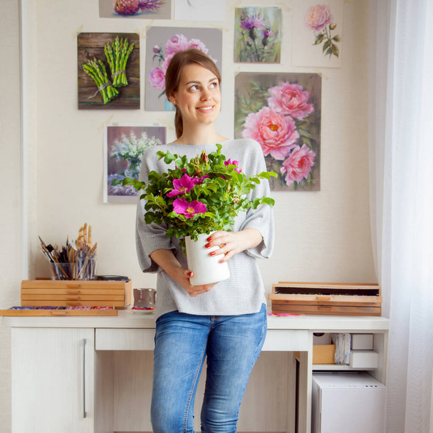 Charming smiling young caucasian woman artist holds a vase in her hands with a beautiful blooming rosehip on the background of her workplace with floral paintings. Art concept - Fotografie, Obrázek