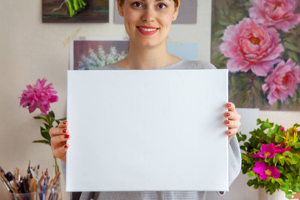 Smiling young woman artist is holding a blank white canvas in her hands against the background of a cozy workplace with paintings hanging on the wall and tools. Advertising space - 写真・画像