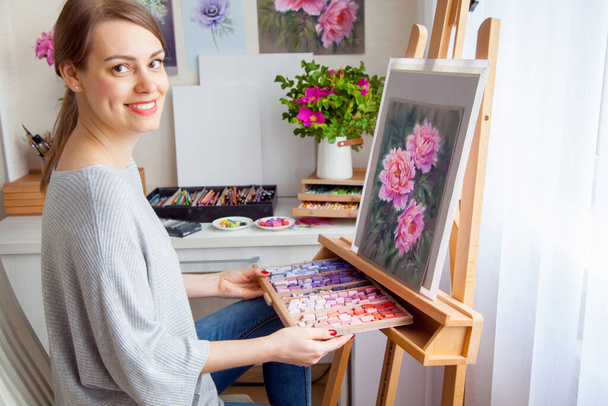 Young smiling caucasian beautiful girl artist paints a picture with pink peonies with the help of crayons dry pastels in a wooden box. Creativity and home hobby concept - Foto, Imagen