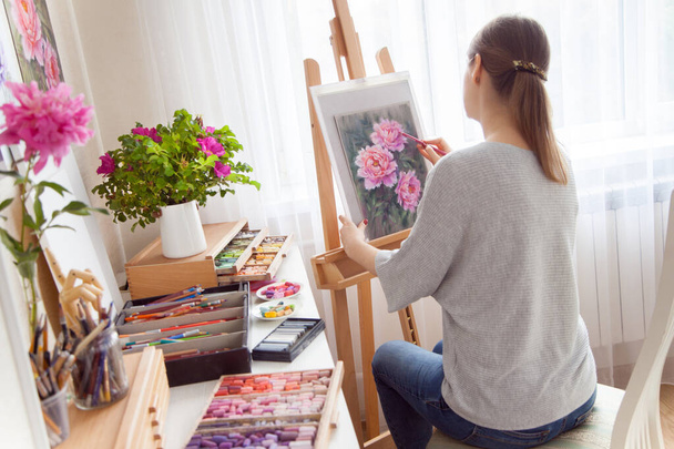 Rear view of young woman artist paints sketch of pink peonies from a box with dry pastels crayons in her studio while sitting at an outdoor wooden easel. Creativity and hobby concept. Pink shades. - Фото, зображення