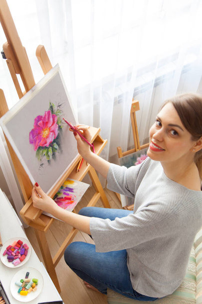 Rear view of young woman artist paints sketch of pink peonies from a box with dry pastels crayons in her studio while sitting at an outdoor wooden easel. Creativity and hobby concept. Pink shades. - Photo, Image