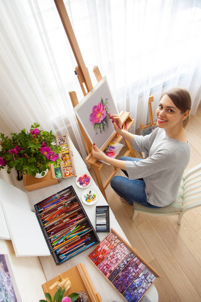 Rear view of young woman artist paints sketch of pink peonies from a box with dry pastels crayons in her studio while sitting at an outdoor wooden easel. Creativity and hobby concept. Pink shades. - Foto, imagen