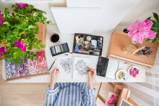 Top view girl artist draws a sketch of rose hip flowers sitting at her workplace with bouquets of flowers and materials for creativity. Hobby and creation concept - Foto, Bild