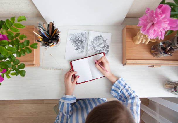 Top view girl artist draws a sketch of rose hip flowers sitting at her workplace with bouquets of flowers and materials for creativity. Hobby and creation concept - Photo, Image