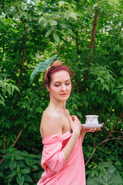 Red hair woman dressed in pink blouse with big green leaf in her hair drinking tea from the white cup in the blooming garden - Fotografie, Obrázek