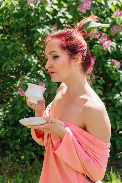 Red hair woman dressed in pink blouse with big green leaf in her hair drinking tea from the white cup in the blooming garden - Photo, Image