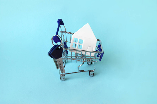Μίνι τρόλεϊ αγορών με λευκό σπίτι και κλειδιά σε μπλε φόντο. Η έννοια της αγοράς ή ενοικίασης ενός σπιτιού, διαμέρισμα, εξοχικό σπίτι και πολλά άλλα. - Φωτογραφία, εικόνα