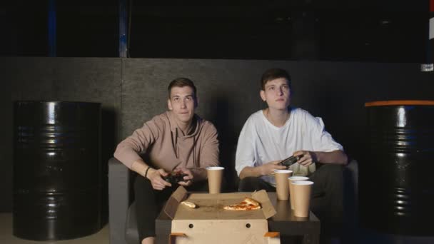 Twee vrolijke vrienden die actie videospel spelen in de woonkamer zittend op de bank. Intense competitie tussen twee jonge mannelijke speelsters zit vol emoties. - Video