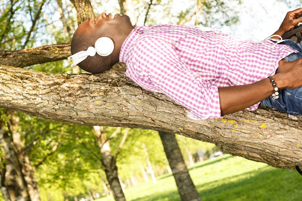 Joven afroamericano acostado en una rama de árbol en un parque y escucha música
 - Foto, imagen
