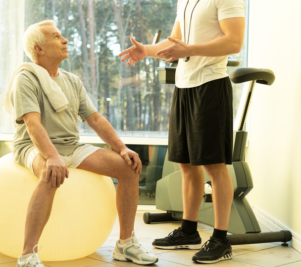 Personal Trainer erklärt einem älteren Mann, wie man mit einem Fitnessball trainiert - Foto, Bild