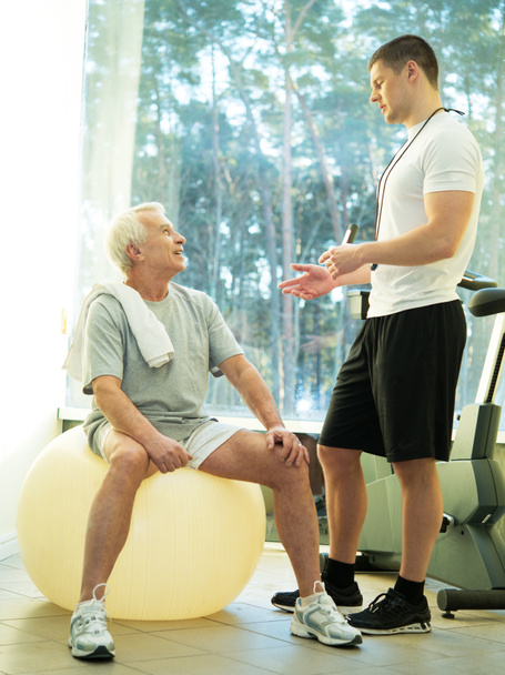 persoonlijke trainer legt een senior man hoe te oefenen op een fitness bal - Foto, afbeelding