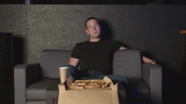 Hombre atractivo comiendo pizza y viendo la televisión en casa usando control remoto relajándose en el sofá - Metraje, vídeo