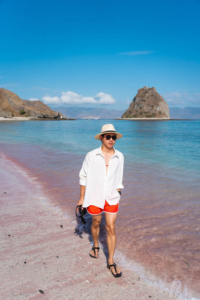 Young Asian man traveler wearing white shirt and walking on pink beach in Komodo national park. A man holding camera and enjoying summer season. Flores island in Indonesia, Asia - Foto, afbeelding