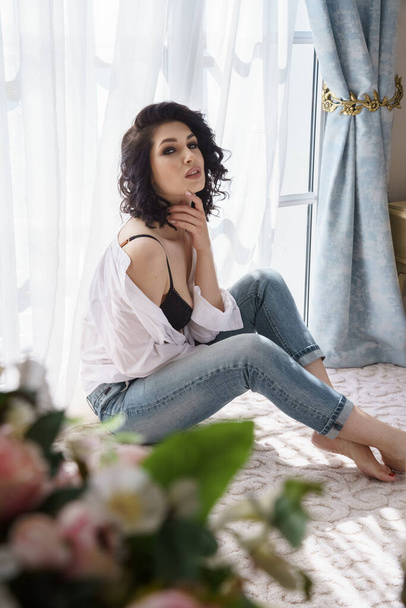 Beautiful curly brunette posing at apartments interior. Portrait of young model in blue jeans and white shirt  - Foto, Imagem
