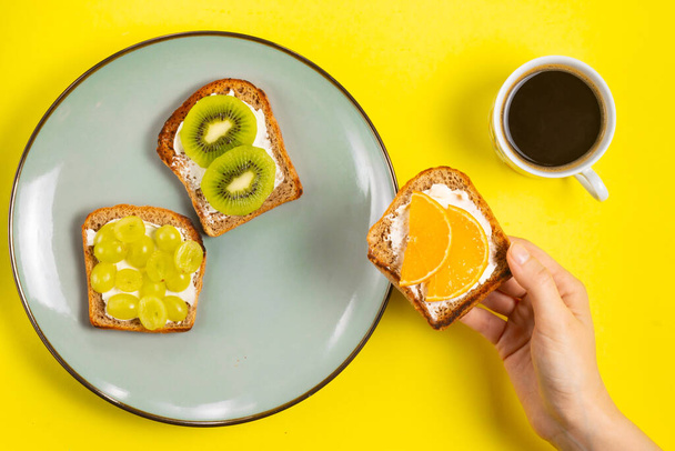 Woman holding in hand a sandwich with cream cheese and orange over a plate with a cup of coffee on a yellow background - Foto, afbeelding
