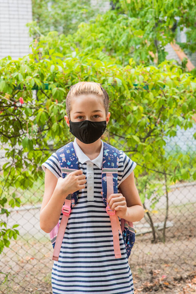 Schoolmeisje meisje met een zwart beschermend masker met een rugzak. Schoolmeisje in schooluniform. Vallen. Binnenkort terug naar school.. - Foto, afbeelding