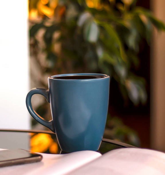 Evening coffee. Blue cup with coffee, open book and mobile phone, evening light, sunset, dusk. Atmospheric, cozy evening in the garden, green leaves, conversation. Warm light, blur bokeh. - Foto, imagen