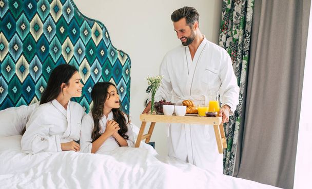 Happy young beautiful family in bathrobes are taking morning breakfast in a luxury hotel room. Service in the room. Vacation and trip - Foto, imagen