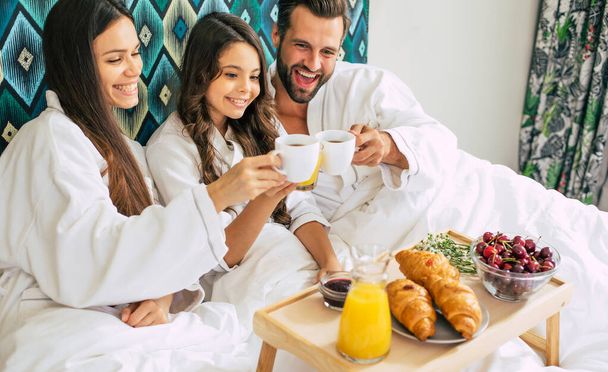 Happy young beautiful family in bathrobes are taking morning breakfast in a luxury hotel room. Service in the room. Vacation and trip - Fotoğraf, Görsel