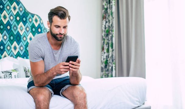 Νέος με αυτοπεποίθηση όμορφος άντρας με μπλουζάκι χρησιμοποιώντας ένα smartphone ενώ κάθεται στο κρεβάτι στο δωμάτιο. Ηλεκτρονική εργασία - Φωτογραφία, εικόνα