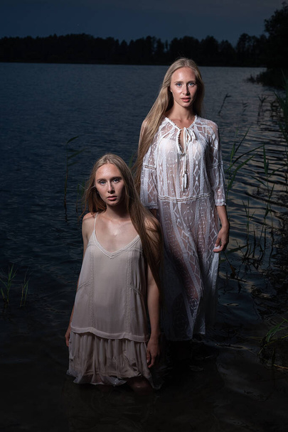 dwie młode siostry bliźniaczki pozujące w jasnych sukienkach w wodzie jeziora w letnią noc - Zdjęcie, obraz