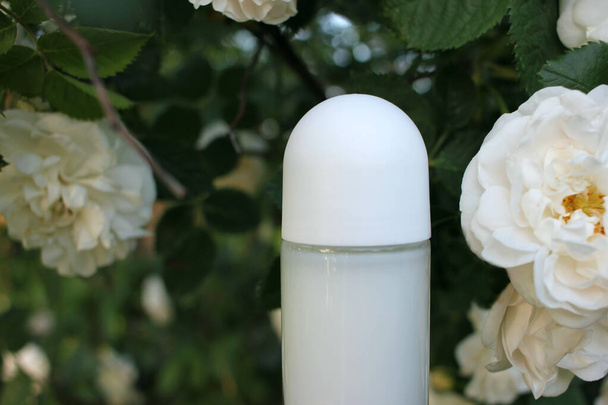 Déodorant antisudorifique roll-on blanc dans la bouteille en verre et roses blanches sur le fond vert naturel flou à l'extérieur - Photo, image