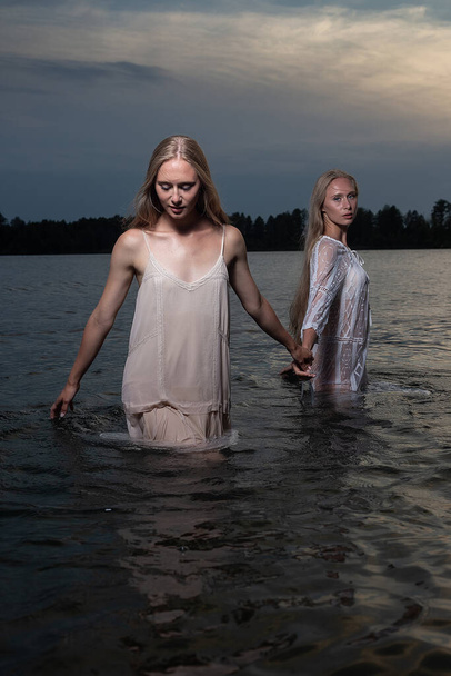 dos hermanas gemelas jóvenes posando en vestidos ligeros en el agua del lago en la noche de verano
 - Foto, imagen