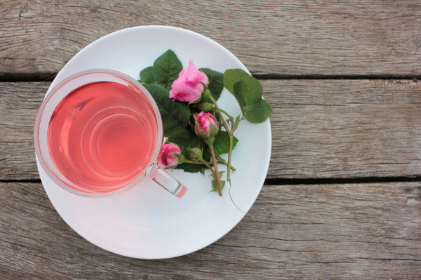 Herbata różana herbata ziołowa w szklanym kubku z różowymi pączkami i kwiatami na rustykalnym drewnianym stole. Letnia herbata na świeżym powietrzu - Zdjęcie, obraz