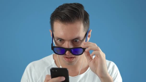 Σοκαρισμένος άνθρωπος με γυαλιά ηλίου Αντιδρά μήνυμα στο Smartphone. Απρόσμενα έκπληκτος και φοβισμένος. Άντρας είναι συγκλονισμένος από αυτό που είδε στο smartphone του, σε μπλε χρώμα φόντο. - Πλάνα, βίντεο