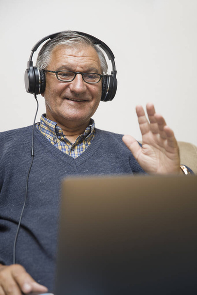 Moderno uomo anziano sito a casa con auricolari e avere videochiamata online - Foto, immagini
