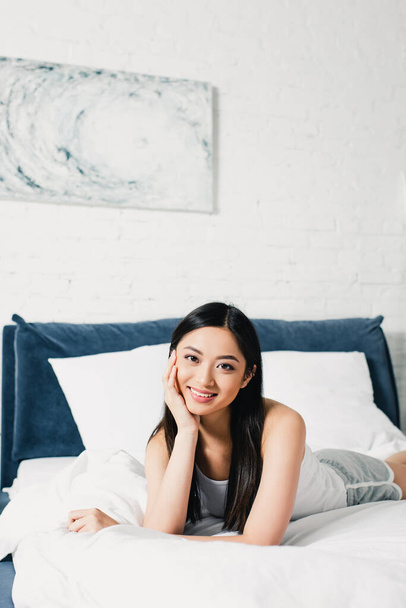 Young asian woman with hand near head smiling at camera on bed  - Photo, Image
