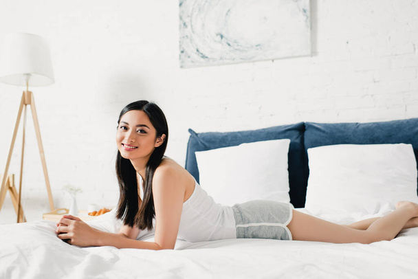 Attractive asian girl smiling at camera while lying on bed  - Valokuva, kuva