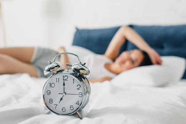 Selective focus of alarm clock on bedding and young woman lying on bed at morning  - Photo, Image