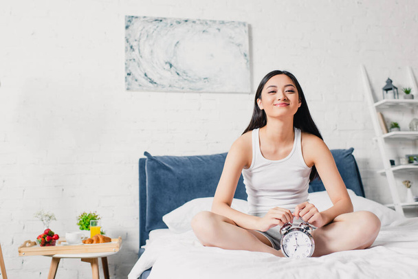 Happy asian woman smiling at camera near alarm clock on bed at morning  - Foto, afbeelding