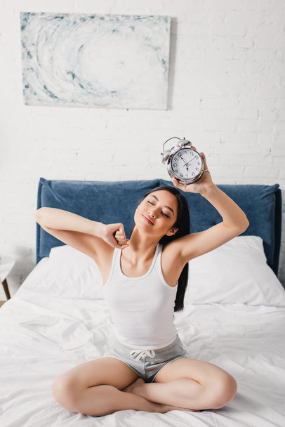 Νεαρή Ασιάτισσα κρατάει ξυπνητήρι και ξαπλώνει στο κρεβάτι το πρωί  - Φωτογραφία, εικόνα